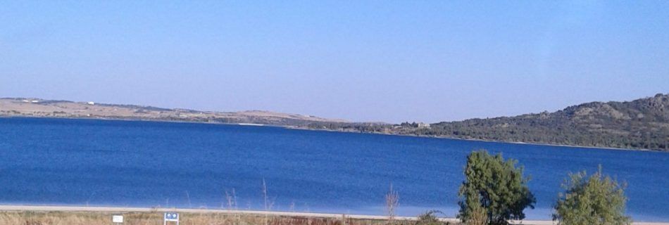 A blue reservoir surrounded by hills and shrubs, under a clear sky.