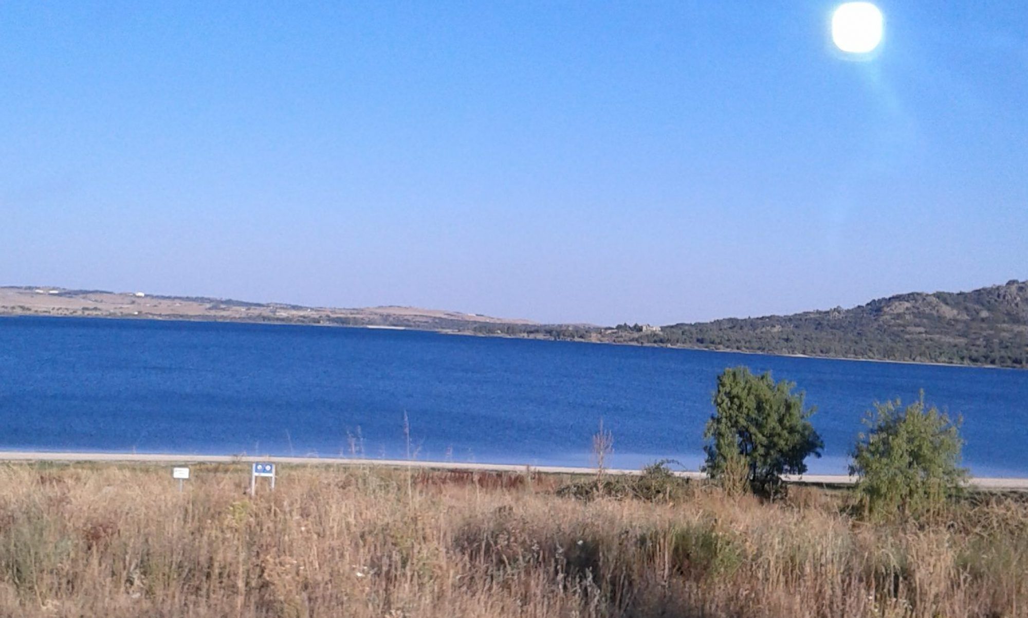 A blue reservoir surrounded by hills and shrubs, under a clear sky.